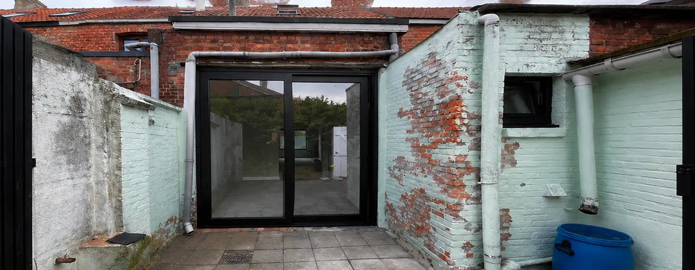 Schuifdeur van zwart omlijst glas, ingebouwd in een verweerde witgeschilderde bakstenen muur met blootliggende rode bakstenen en leidingen.